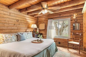 A wooden cabin bedroom with a bed, a chair, a window with curtains, a ceiling fan, and a nightstand with a lamp. - Doe Creek Ranch | 2BR Cabin with Horse Amenities (Bandera)