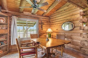 A wooden cabin interior with a dining table, chairs, a ceiling fan, a window with curtains, and a mirror on the wall.
