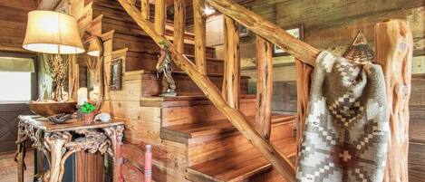 A wooden staircase with a handrail, a table with a lamp, and a woven chair.