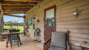 A rustic wooden cabin with a porch, a dining table with chairs, a fan, and a cushioned chair. - Doe Creek Ranch | 2BR Cabin with Horse Amenities (Bandera)