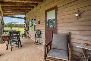 A rustic wooden cabin with a porch, a dining table with chairs, a fan, and a cushioned chair. - Doe Creek Ranch | 2BR Cabin with Horse Amenities (Bandera)
