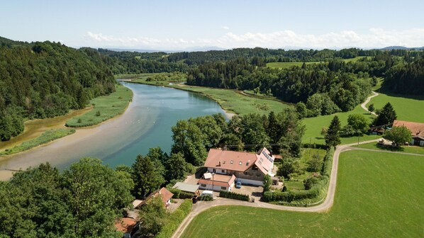 A rural house with a red-tiled roof, surrounded by lush greenery and a river.