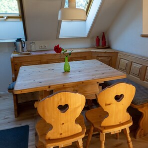 A wooden dining table with a green vase and a red rose. There are two wooden chairs with heart-shaped cutouts. A skylight is above the table, and a window is visible to the left. - Idyllic vacation apartment with 2 bedrooms and balcony in the house on the Iller (Altusried)