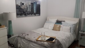 A neatly made bed with a tray of bottled water and a smartphone, a cityscape painting on the wall, and a lamp on a bedside table. - The Jahmtons Guest Room (Ridge)