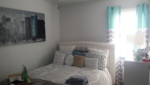 A neatly made bed with a white headboard, a cityscape painting on the wall, and a window with teal curtains. - The Jahmtons Guest Room (Ridge)