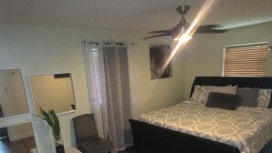 A bedroom with a bed, a chair, a small table with a plant, a ceiling fan, and a window with blinds.