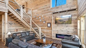 Interior acogedor de una cabaña de madera con chimenea, una escalera y un gran ventanal con vistas a un bosque.