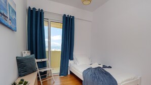 Un dormitorio con cama, una silla, una mesita con plantas y vistas al exterior a través de una ventana con cortinas.