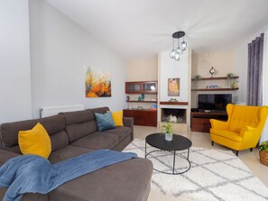 A modern living room with a sofa, armchair, and coffee table. There is a fireplace, a TV, and a bookshelf.