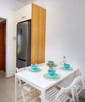 A modern kitchen with a stainless steel refrigerator, wooden panel, white cabinets, and a white dining table with blue plates and a small potted plant.