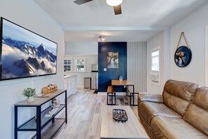 A modern living room with a leather sofa, a wooden coffee table, a TV mounted on the wall, and a kitchen area in the background.