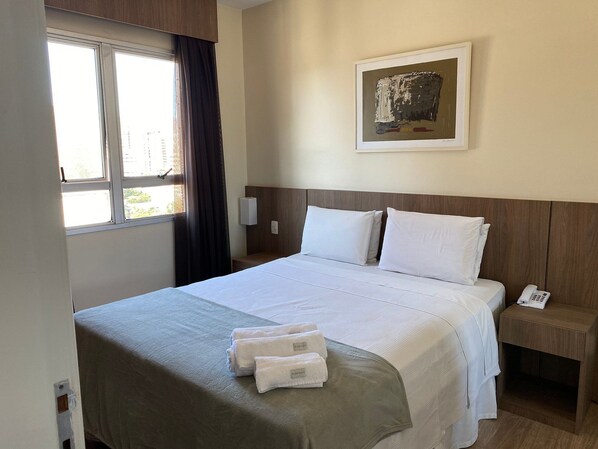A hotel room with a bed, a bedside table with a phone, a window with curtains, and a painting on the wall.