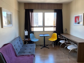Un espacio habitable compacto con sofá, una mesita, dos sillas, un escritorio y un ventanal con cortinas.