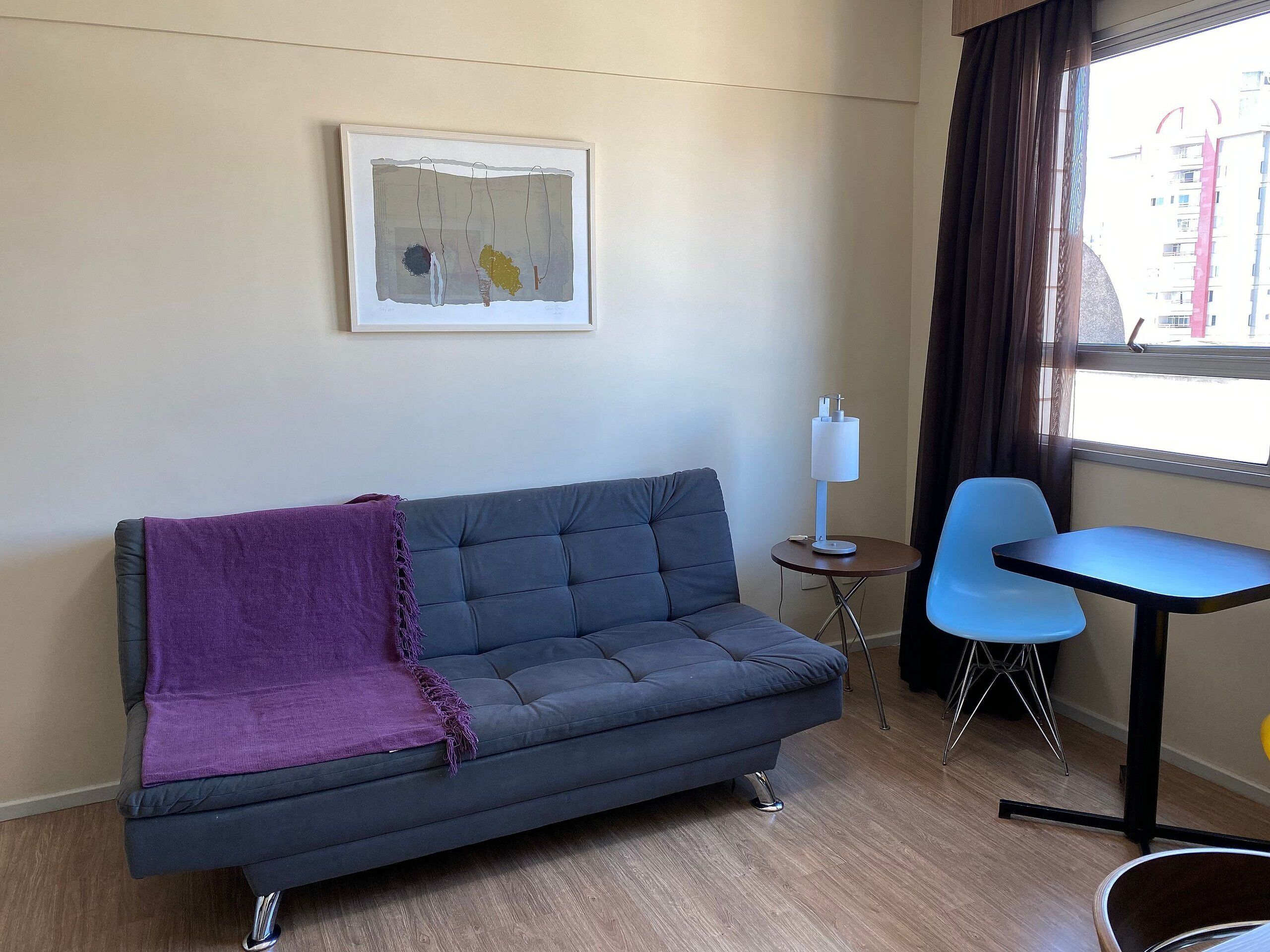 A modern living room with a grey sofa, a purple throw blanket, a small round table with a lamp, and a blue chair.