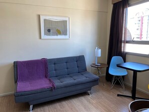 A modern living room with a grey sofa, a purple throw blanket, a small round table with a lamp, and a blue chair.