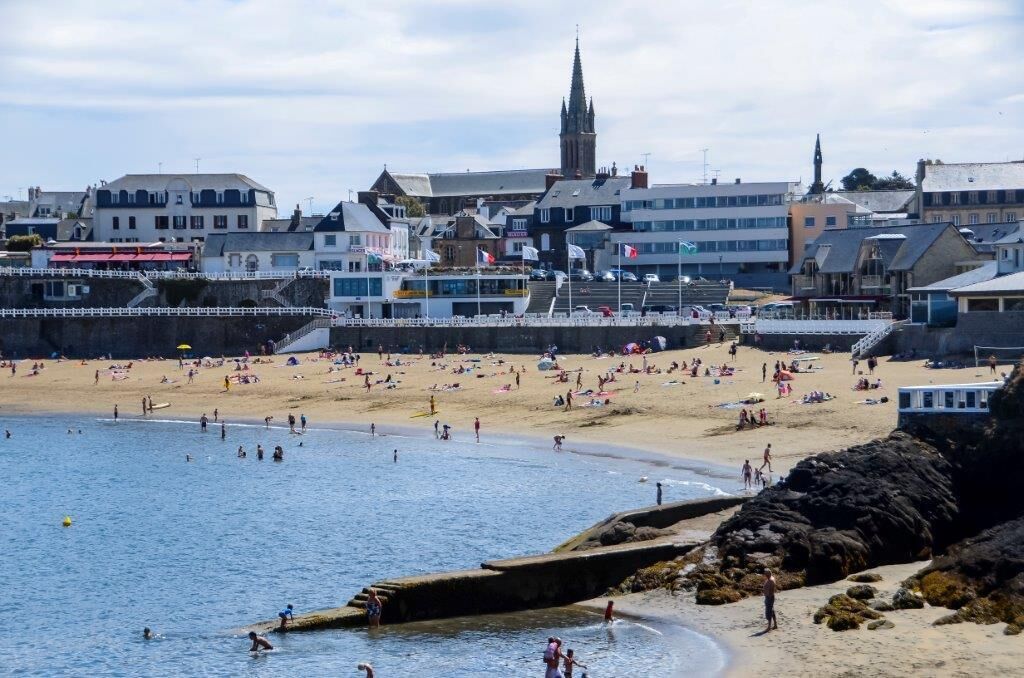 A beach with people sunbathing and swimming, a pier, and buildings in the background.