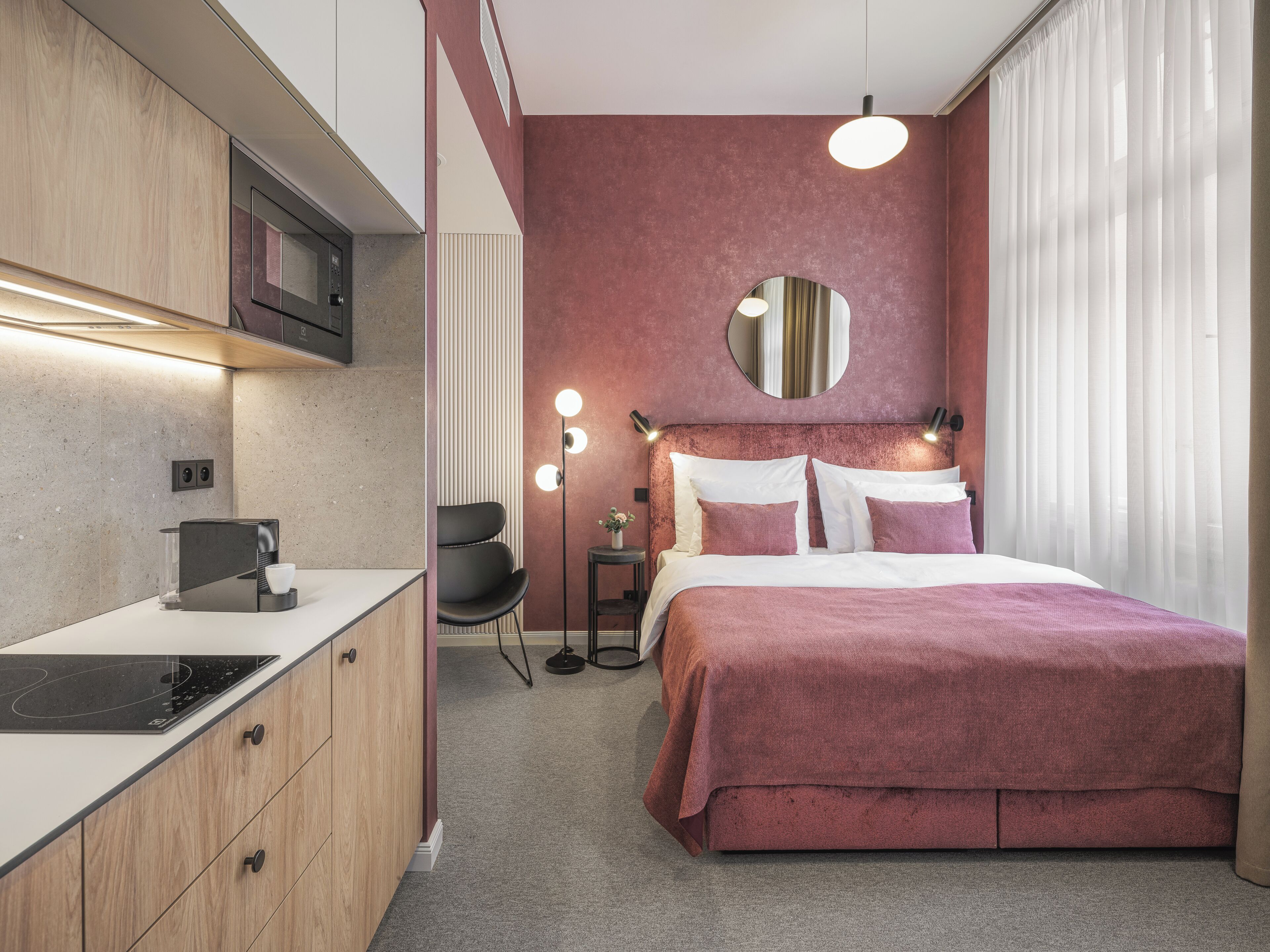 A compact hotel room with a kitchenette, a bed with a pink bedspread, a round mirror above the bed, and a small side table with a lamp.