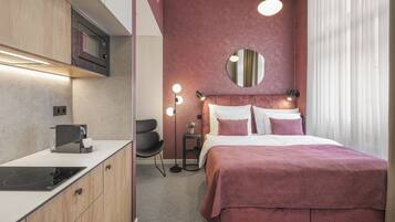 A compact hotel room with a kitchenette, a bed with a pink bedspread, a round mirror above the bed, and a small side table with a lamp.