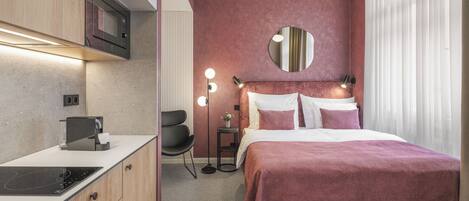 A compact hotel room with a kitchenette, a bed with a pink bedspread, a round mirror above the bed, and a small side table with a lamp.