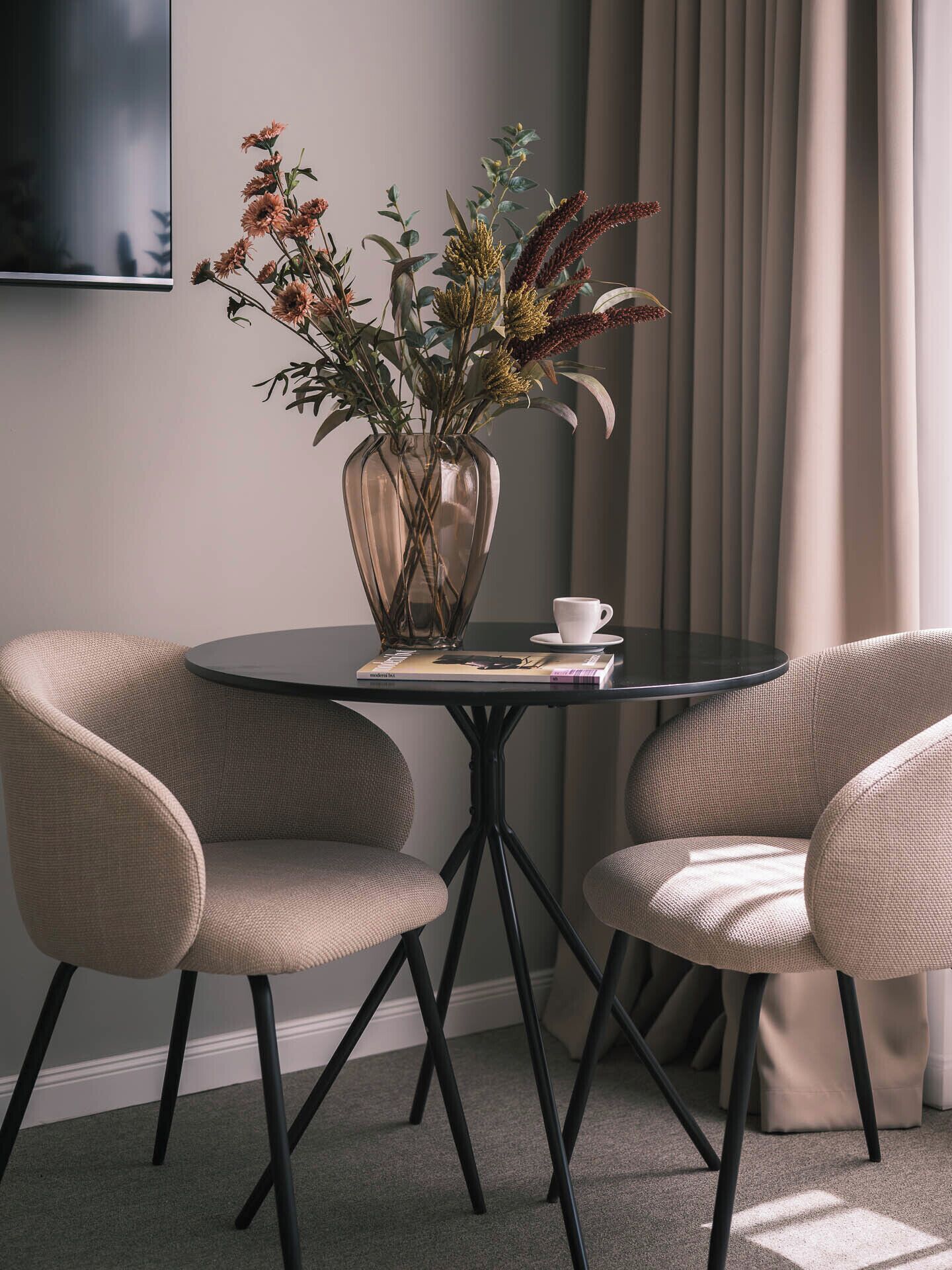 A modern room with a round table, two chairs, a vase with dried flowers, and a remote control on the table.