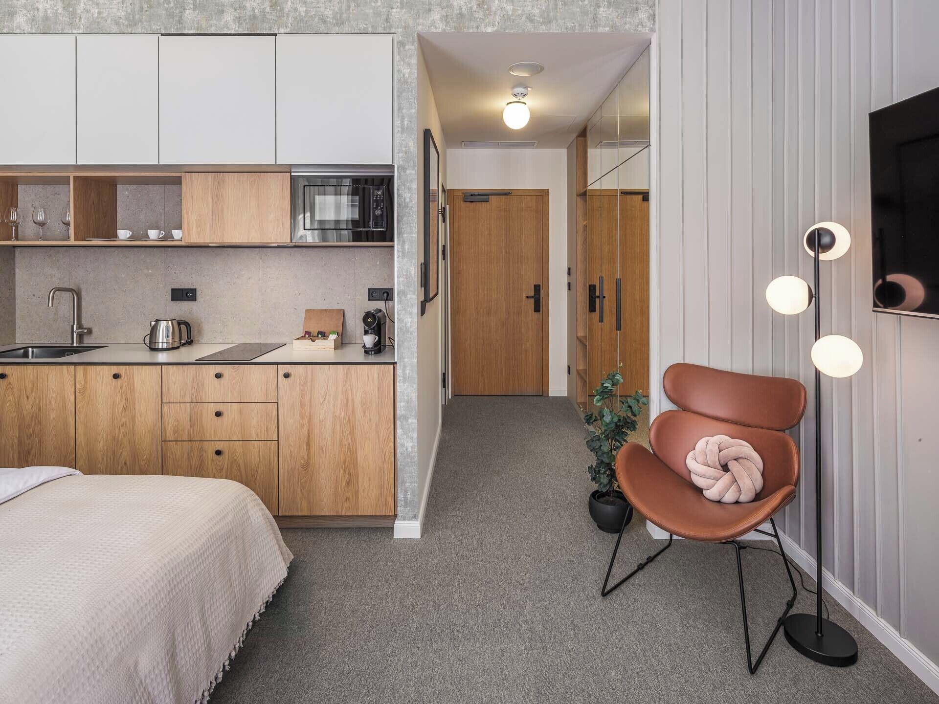 A compact hotel room with a kitchenette, a bed, a seating area with a chair, and a potted plant.