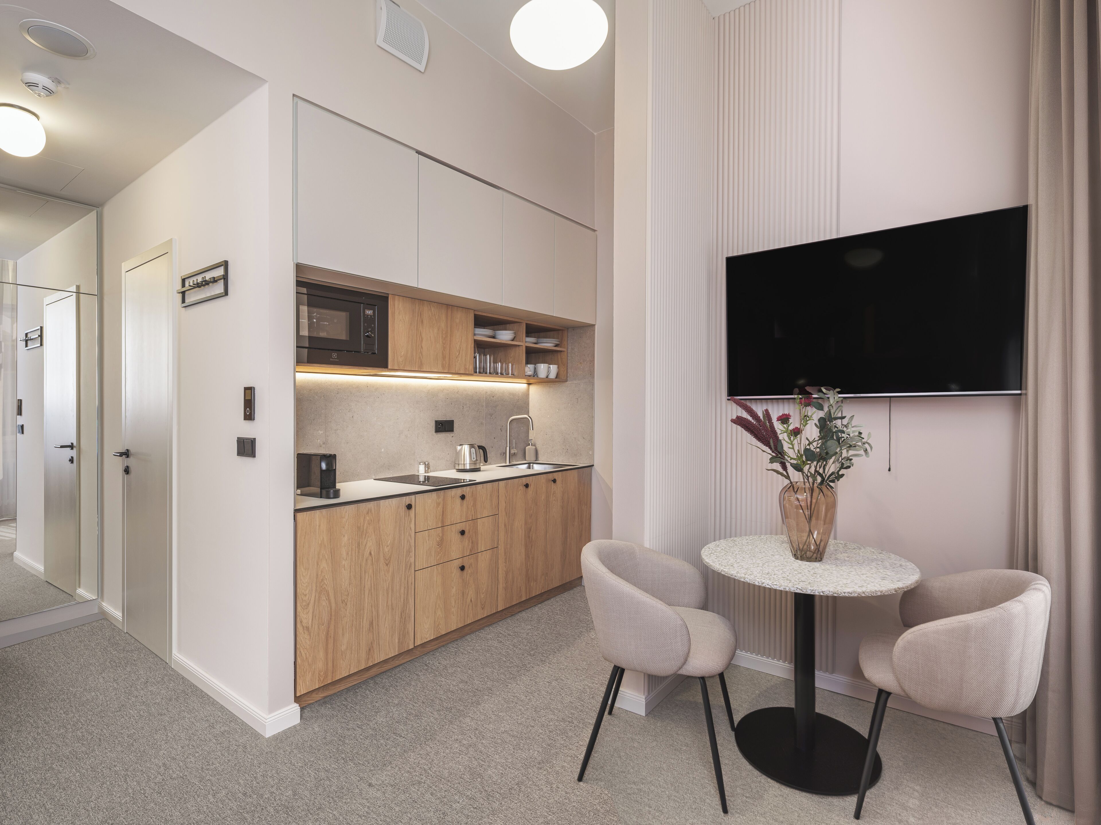 A modern kitchen with wooden cabinets, a built-in microwave, and a coffee maker. There is a round table with two chairs and a vase with flowers.