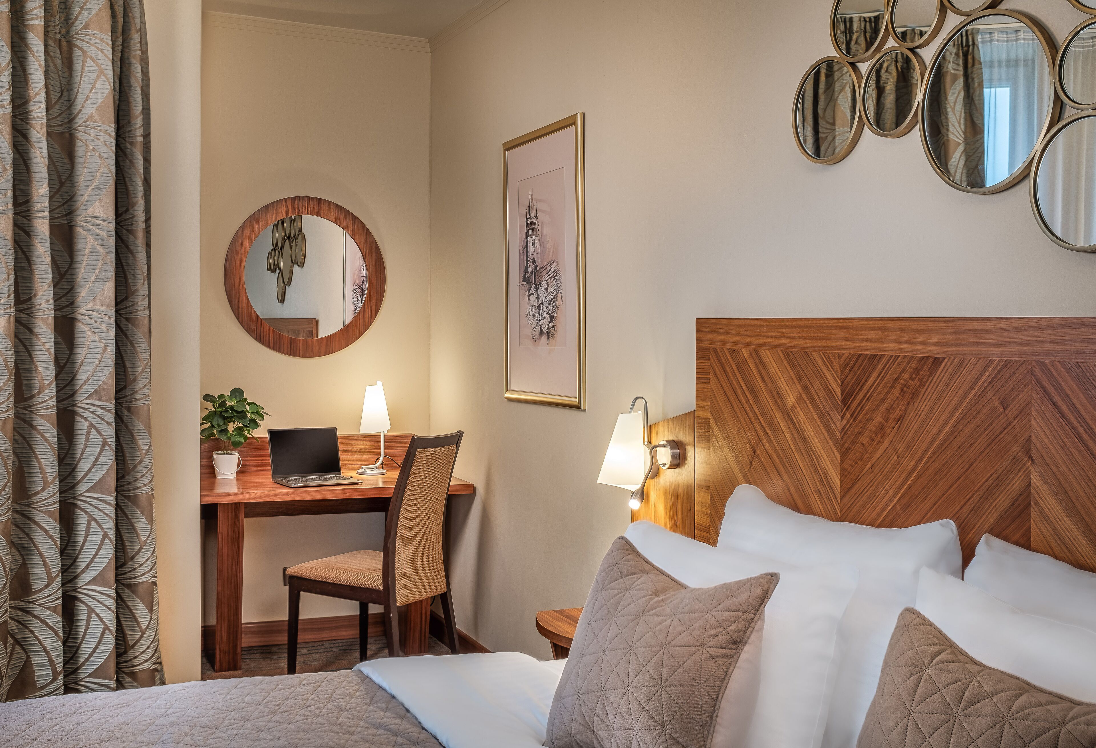 Habitación de hotel con cama, escritorio con computadora portátil, silla, lámpara y espejo en la pared.