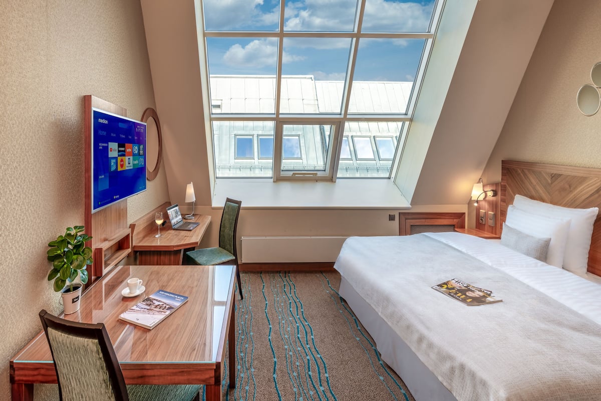 A hotel room with a large window, a desk with a laptop, a chair, a bed with pillows, and a wall-mounted TV.