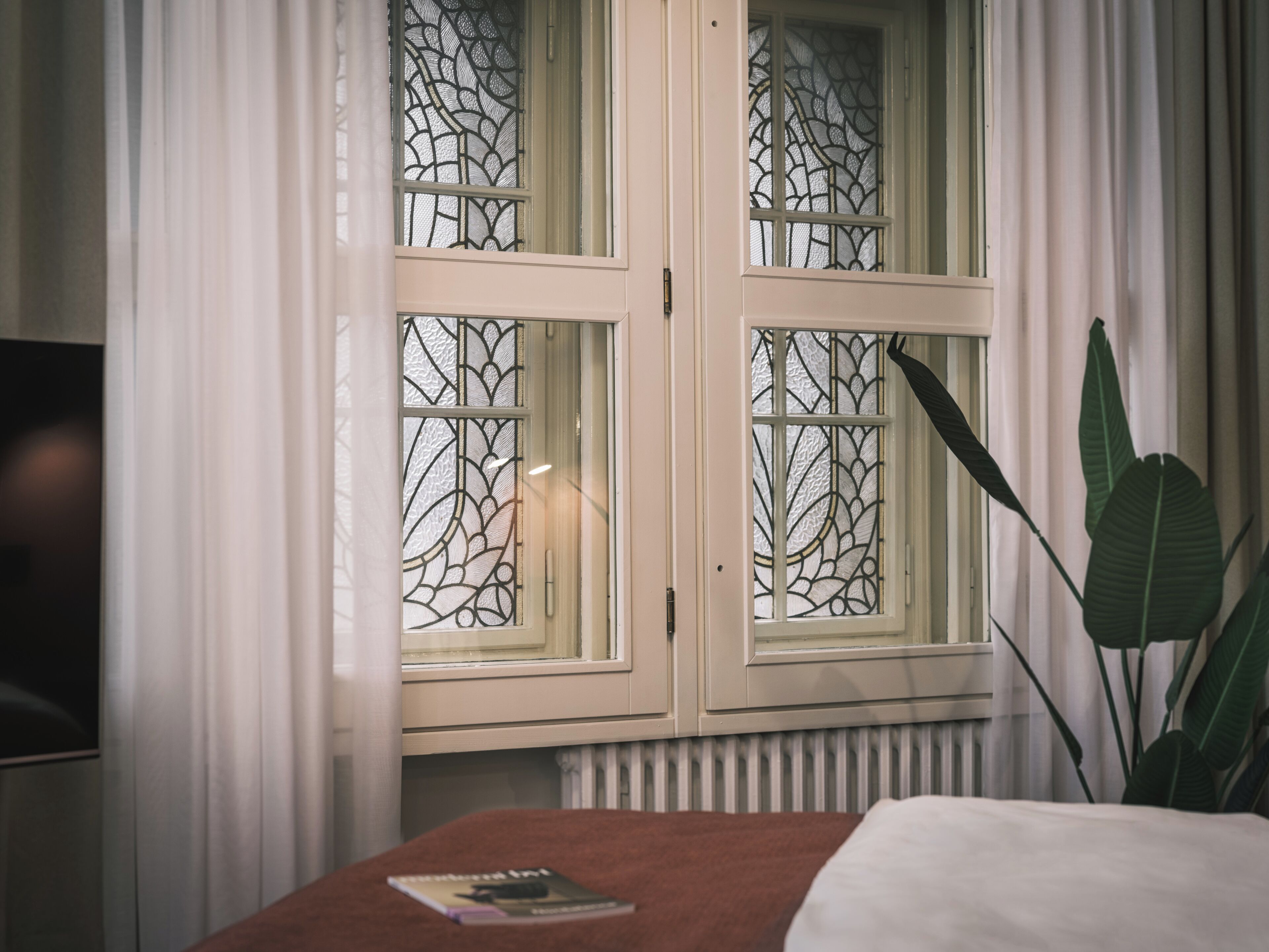Een kamer met een bed, een raam met decoratief glas, een radiator en een plant.
