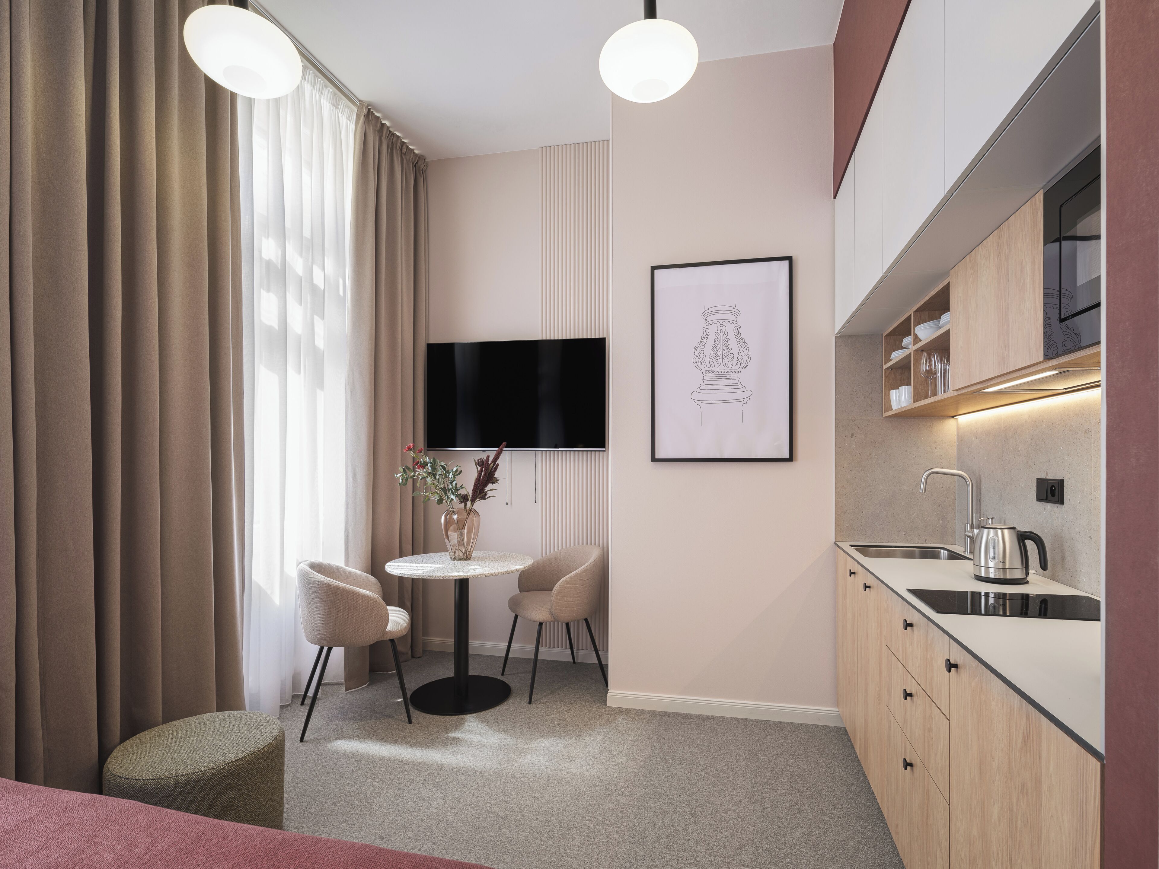 A modern hotel room with a kitchenette, a round dining table with chairs, a flat-screen TV mounted on the wall, and a framed artwork above the TV.