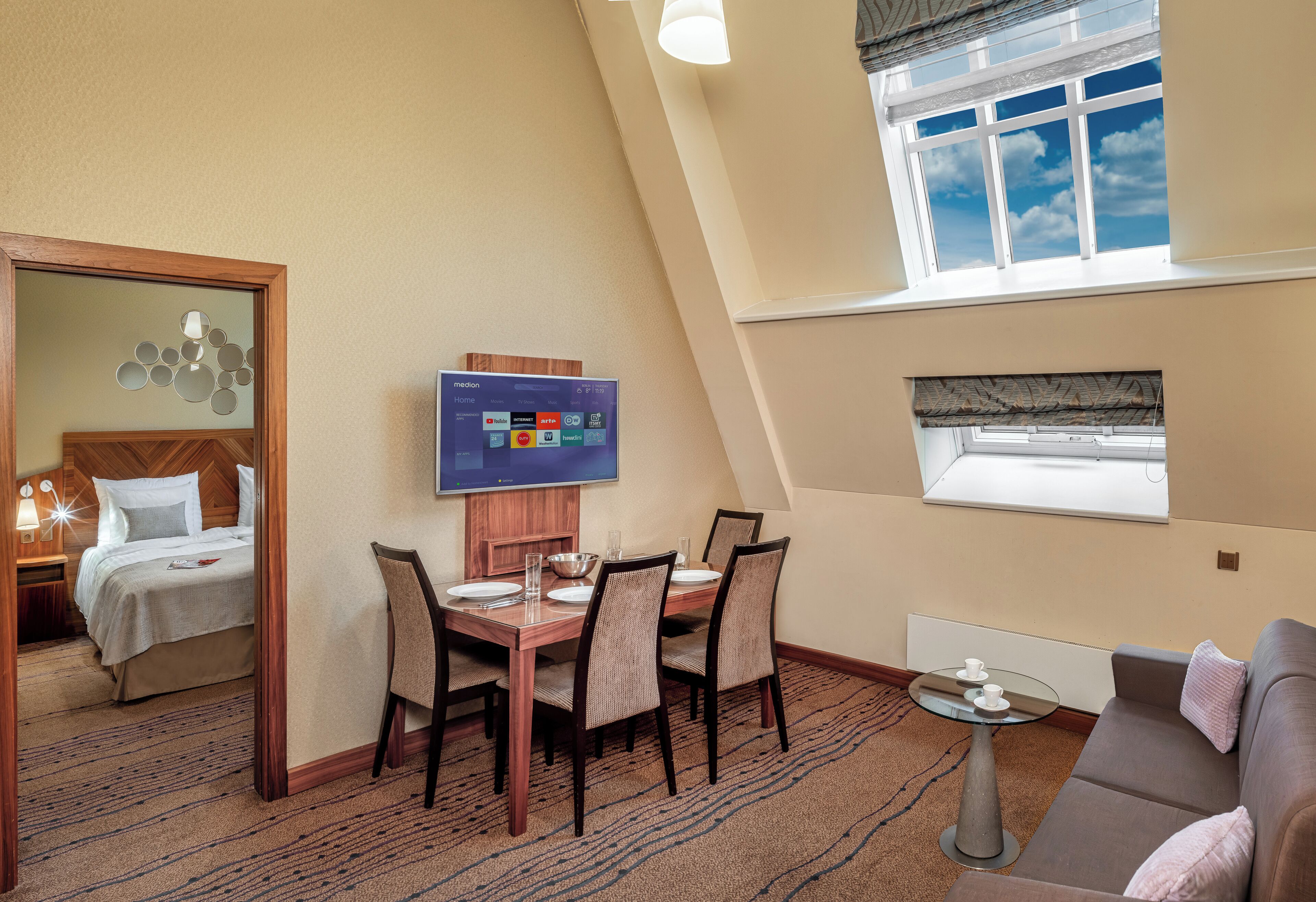 A hotel room with a dining area, a TV mounted on the wall, a sofa, and a window with a view of the sky.