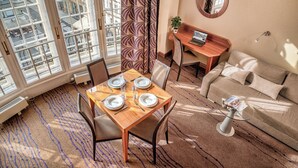Un coin repas avec une table dressée pour quatre personnes, un canapé, une petite table ronde avec un ordinateur portable, et une grande fenêtre donnant sur des immeubles.