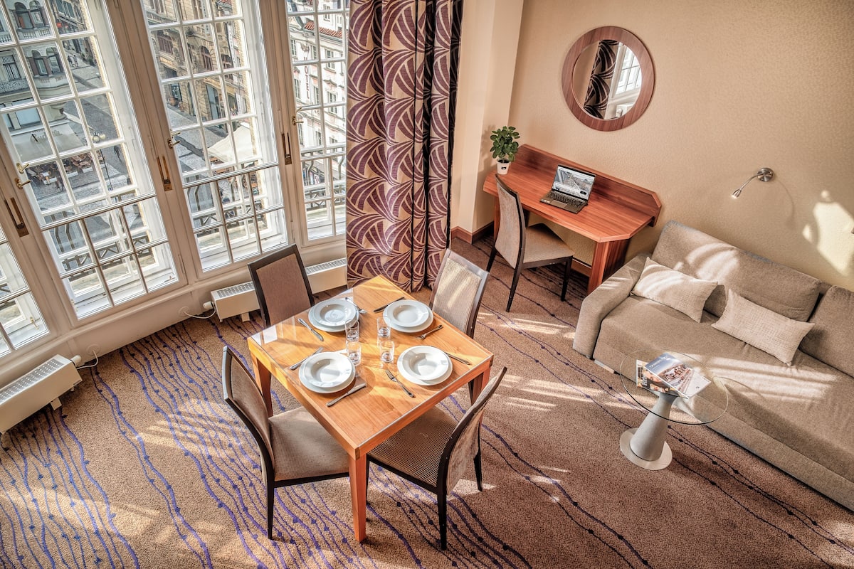 A dining area with a table set for four, a sofa, a small round table with a laptop, and a large window with a view of buildings.