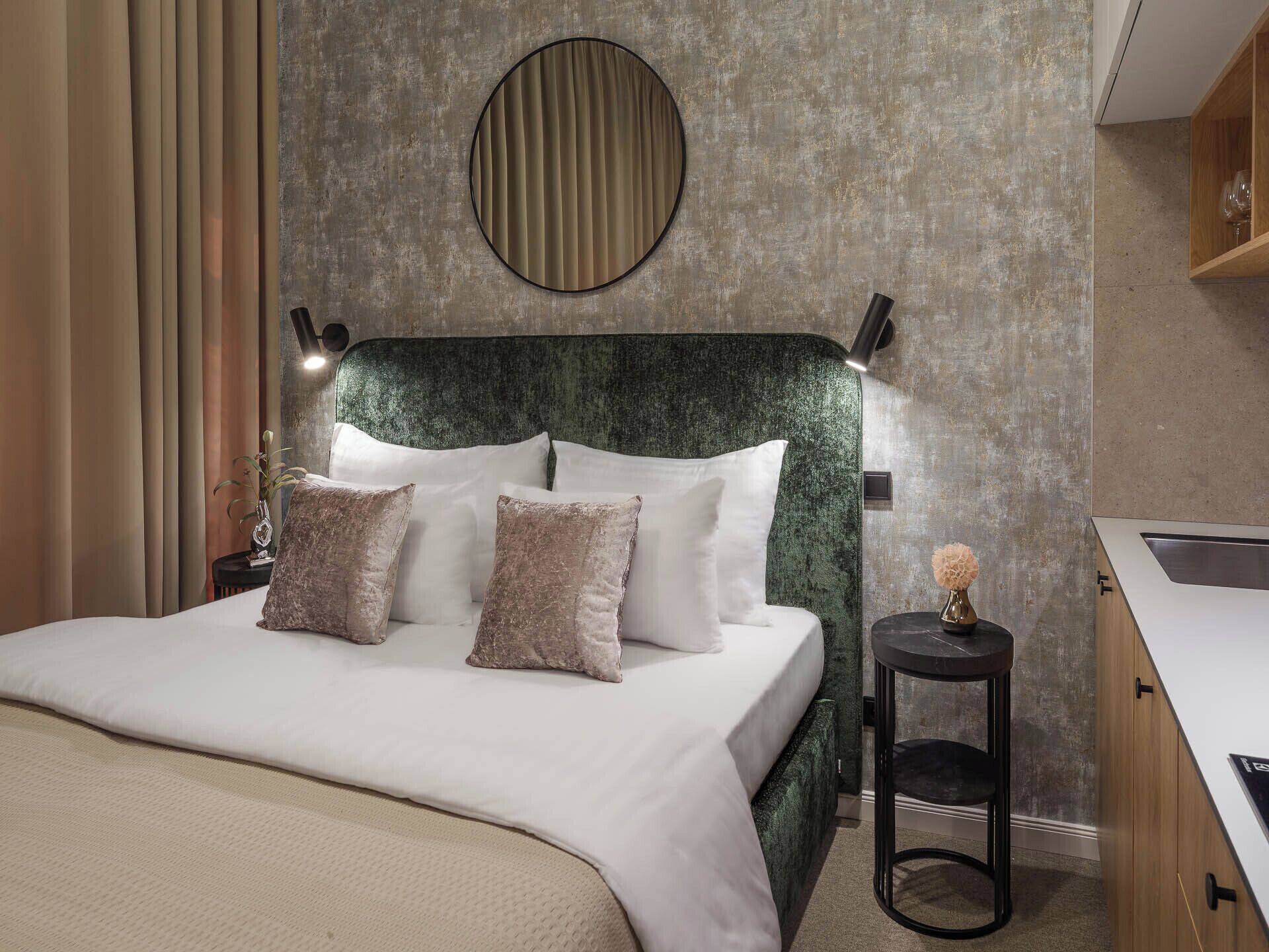 Habitación de hotel con una cama, un espejo redondo, una mesita con un florero y una zona de cocina integrada.