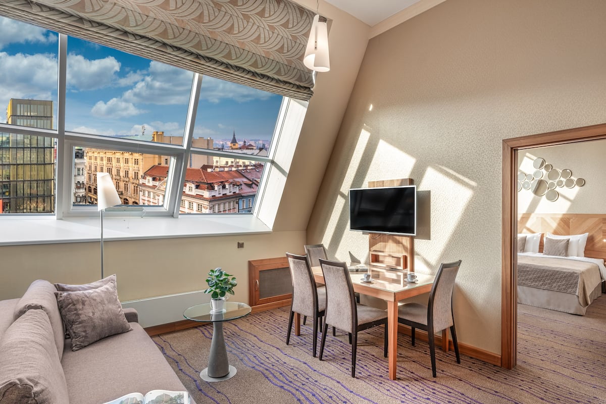A hotel room with a large window offering a city view, a cozy seating area with a sofa and a small table, a desk with a computer and chair, and a bed with a wooden headboard.