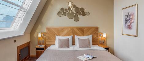 A hotel room with a sloped ceiling, a wooden headboard, two bedside tables with lamps, a large bed with a quilted cover, and a window with a view of the sky.