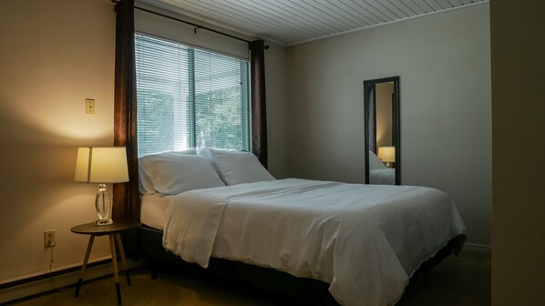 A neatly made bed in a hotel room with a bedside lamp, a mirror, and a window with blinds.