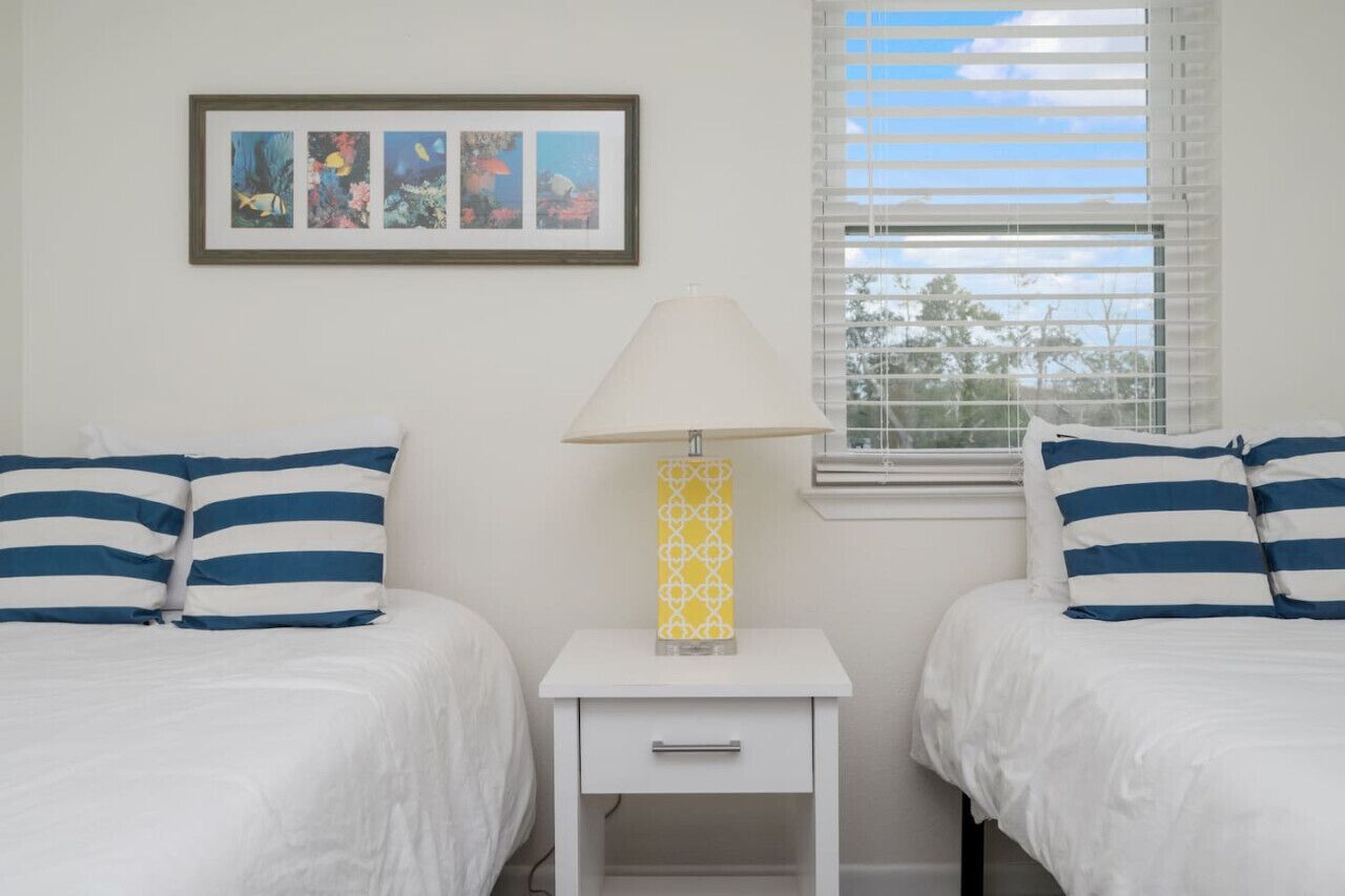 A bedroom with two beds, a bedside table with a lamp, and a window with blinds.