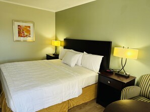 A neatly made bed with white linens, a dark headboard, two bedside lamps, a small bedside table, and a striped armchair. - Astoria North Extended Stay (Danville)