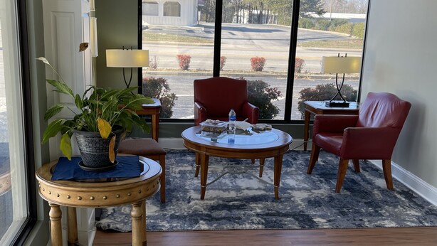 A spacious living room with a large window, a round coffee table, two red chairs, and a potted plant. - Astoria North Extended Stay (Danville)