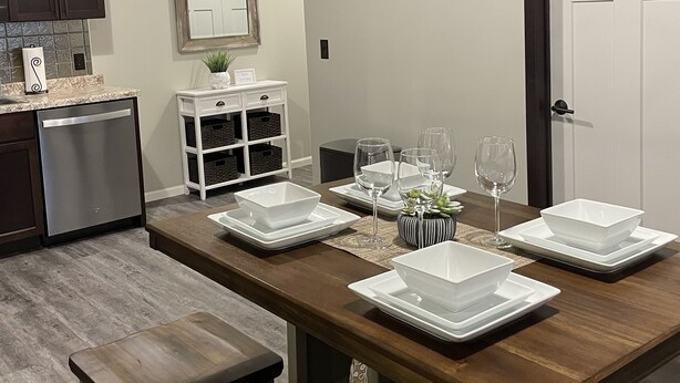 Un comedor con una mesa de madera dispuesta para seis personas, platos blancos y copas de vino. Hay dos taburetes en la mesa, un armario blanco con estantes, y un mueble de madera oscura.