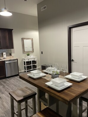 A dining area with a wooden table set for six, white plates, and wine glasses. There are two stools at the table, a white cabinet with shelves, and a dark wooden cabinet.