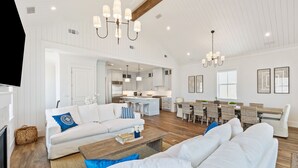 A spacious living area with a white sofa set, a coffee table, and a dining table with chairs. A kitchen is visible in the background.