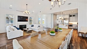 A spacious living room with a large TV, white sofas, a wooden dining table, and a kitchen area visible through an open doorway.