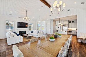 A spacious living room with a large TV, white sofas, a wooden dining table, and a kitchen area visible through an open doorway.