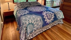 Um quarto com teto de madeira, duas janelas com persianas, uma cama com colcha estampada, duas luminárias de cabeceira e vista para o verde lá fora.