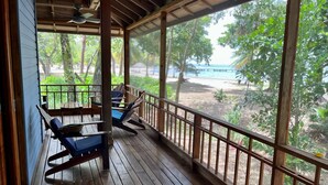 Um deck de madeira com duas cadeiras de descanso e um ventilador de teto, com vista para uma praia e vegetação exuberante.