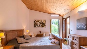 Une chambre à coucher avec une tête de lit en bois, un lit recouvert d’une couvre-lit beige, une table de chevet avec une lampe, une petite table avec un vase de fleurs et une commode en bois.
