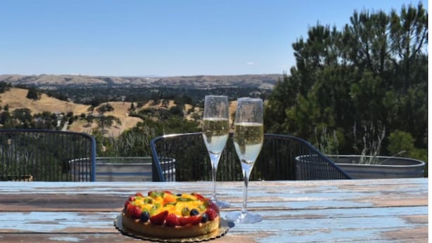 Une table avec une tarte aux fruits et deux verres de vin blanc, le tout mis en valeur par un paysage pittoresque de collines et d’arbres.