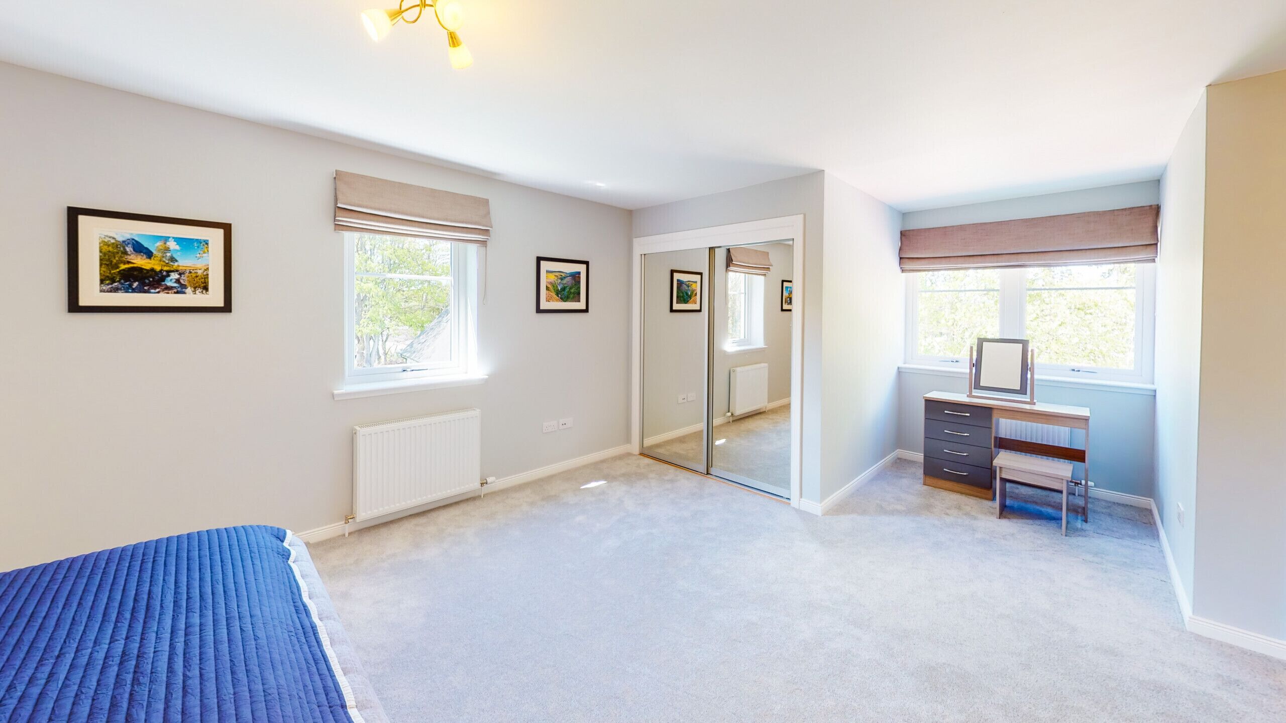 A bedroom with a bed, a desk with a computer, a mirror, and two windows with blinds.